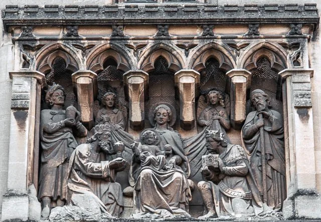 A detailed stone relief sculpture depicting a religious scene with multiple figures, including a central seated woman holding a child, surrounded by other seated and standing figures with various objects, set within an arched structure.