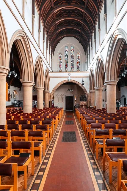 Interior of a church with rows of wooden pews, stained glass windows, and wooden ceiling beams.