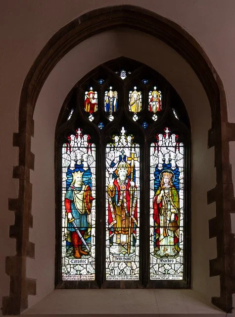 Colorful stained glass window featuring three central figures and smaller figures above, with labels 'Caradoc,' 'Dewi Sant,' and 'Maer.