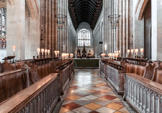 Inside a historic church with high vaulted ceilings, stone walls, wooden pews on either side, stained glass windows, and candles on small tables along the pews.