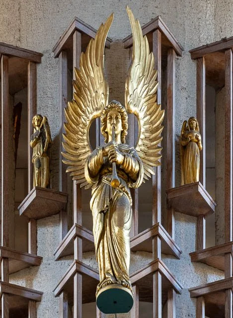 Golden angel sculpture with large wings holding a sword, flanked by two smaller gold figures, displayed on a church wall.