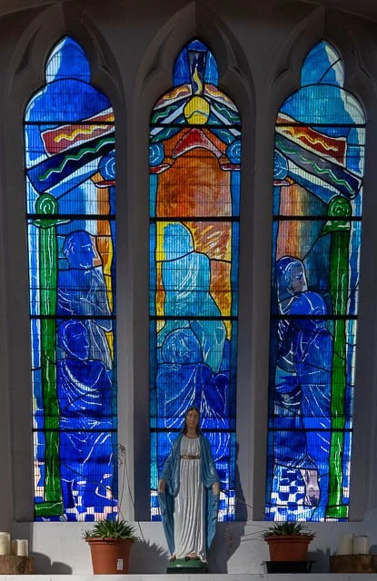 Colorful stained glass window depicting religious figures and a sun with rays, with a statue of Jesus Christ in front.