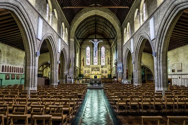 Interior of a church with rows of wooden pews, arched stone walls, stained glass windows, and an altar with a crucifix at the front.