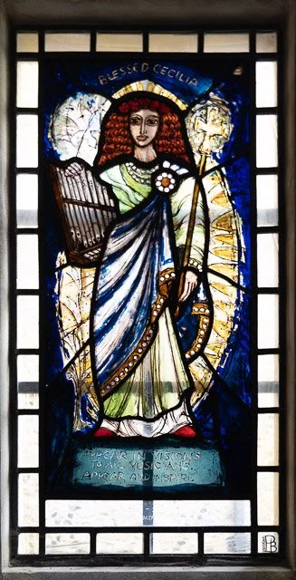 Stained glass window of a saint, holding a harp and a staff, with a halo and a crown of red roses, surrounded by clouds and blue sky, with the name Bless'd Cecilia at the top.