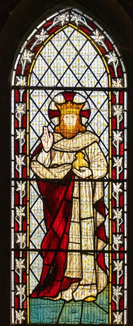 Stained glass window depicting a crowned figure of Jesus Christ holding a cup, with a red and white robe, and making a blessing gesture, surrounded by geometric and floral patterns