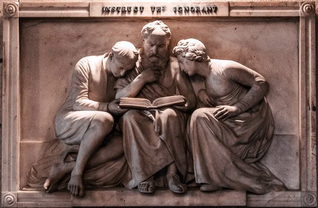 A stone sculpture of two women reading a book with Jesus in the middle, with the words "Instructed by Ignorance" inscribed above.