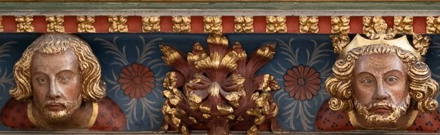Close-up of an ornate carved and painted wooden decorative panel with two faces flanking a central floral design