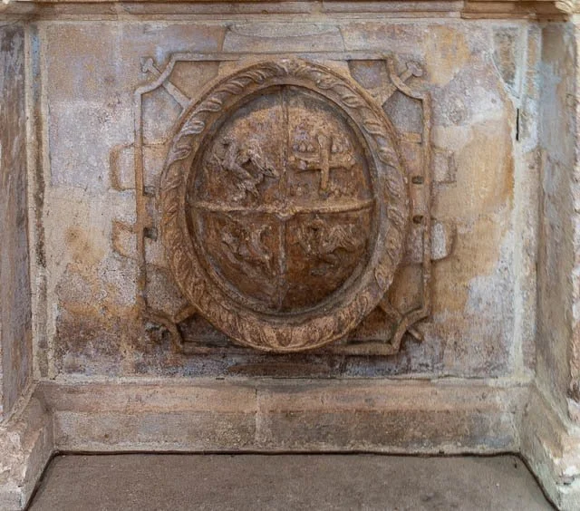 A weathered stone wall with an oval emblem featuring a ship, a cross, and a lion, surrounded by decorative stonework.