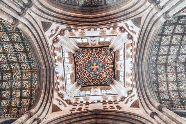 View of an ornate church ceiling with a square-shaped central decorative element surrounded by detailed architecture and stained glass windows.