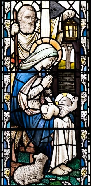 A stained glass window depicting Mary holding baby Jesus, with a shepherd and a lamb nearby, in a biblical scene.