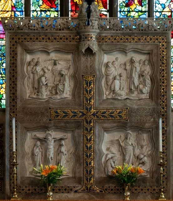Stone religious relief sculpture divided into four sections with a decorative cross in the center, depicting scenes from the Passion of Christ, with stained glass windows in the background.