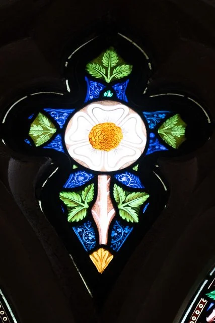 Colorful stained glass window with a white flower with a yellow center surrounded by green leaves and intricate blue and gold decorative details.