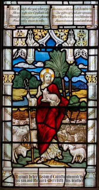 Stained glass window depicting Jesus Christ with a lamb in his arms, sitting outdoors with sheep at his feet, surrounded by ornate architectural details and biblical text.