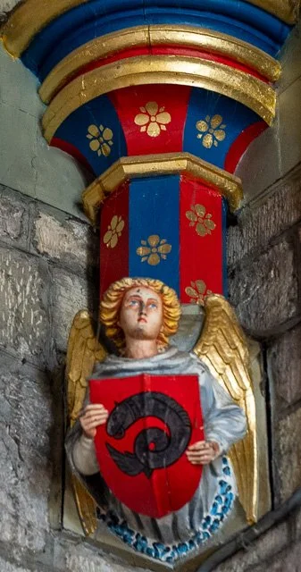 Colorful religious statue of angel with wings holding shield, situated in a stone alcove with decorative blue, red, and gold accents.