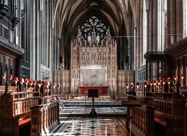 Interior of a Gothic-style church or cathedral with a high vaulted ceiling, ornate wooden balconies, red lamps, and an altar at the front.