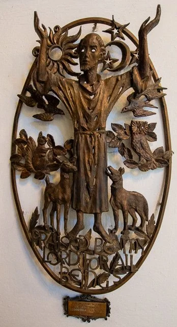 Wooden wall hanging of a human figure with outstretched arms, surrounded by birds, deer, leaves, a sun, and moon, with the words 'Religiosa Corporal' engraved at the bottom.
