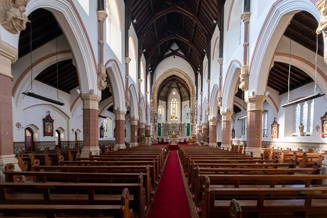 Interior of a church with wooden pews, arched columns, and stained glass windows at the front.