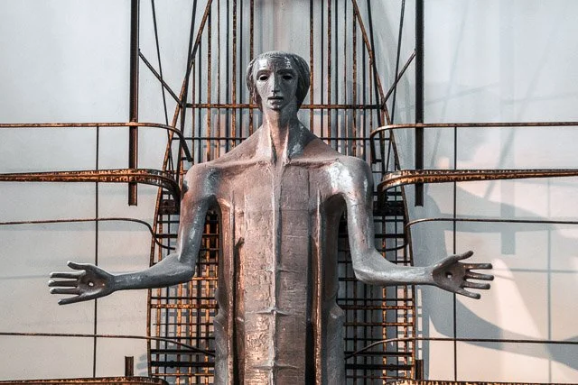 A sculpture of a humanoid figure with an elongated neck and arms outstretched, set against a backdrop of rusty metal framework.