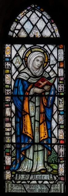 Stained glass window depicting a woman in a blue and orange robe holding a red book, with a halo around her head, framed by decorative patterns.