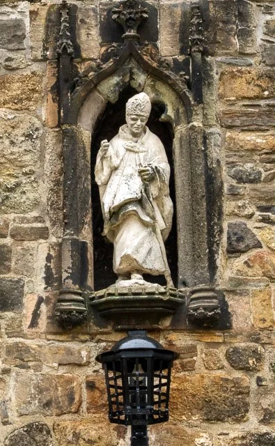 Stone statue of a man in historical clothing inside a niche on a brick wall, with a black lantern below.