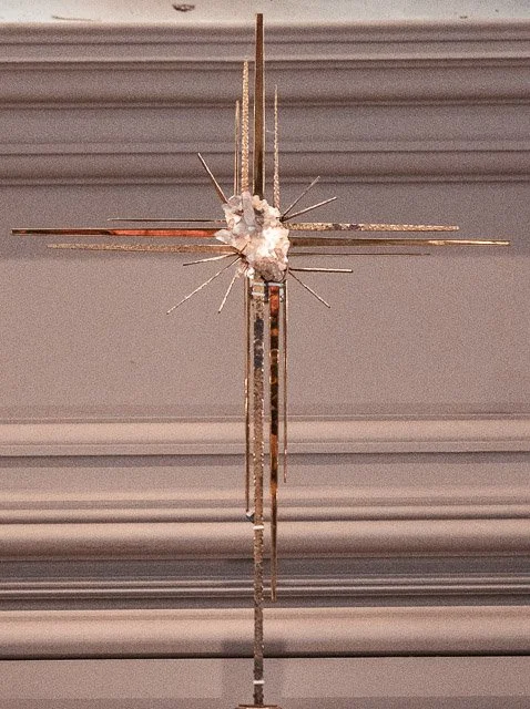 Decorative cross with a central floral arrangement and radiating metal rods, mounted on a wall.