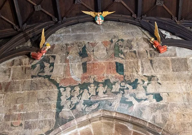 Faded medieval wall painting depicting a group of people gathered around a central figure, with decorative angels or cherubs in a church interior.
