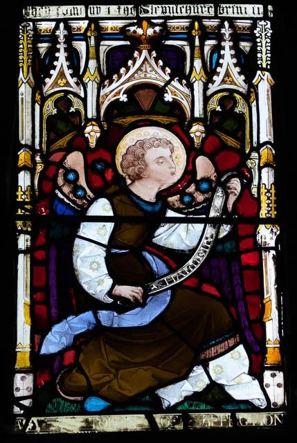 Stained glass window depicting a kneeling angel with curly hair, surrounded by intricate Gothic architectural designs.