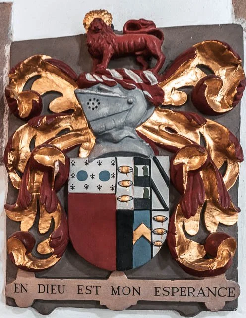 A coat of arms with gold and red decorative elements, a gray eagle, and a red lion figure on top. The shield is divided into sections with blue and white patterns, red, black, and blue shapes. There is a Latin motto at the bottom: "EN DIEU EST MON ES