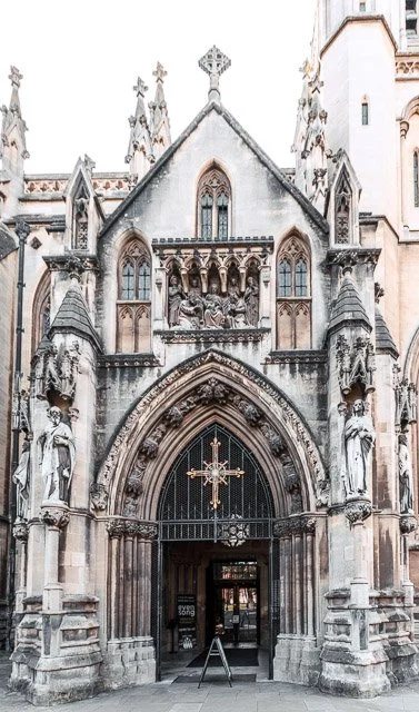 Front view of a Gothic-style church or cathedral with detailed stone carvings, statues, and a large arched doorway with a cross and decorative elements above the entrance.