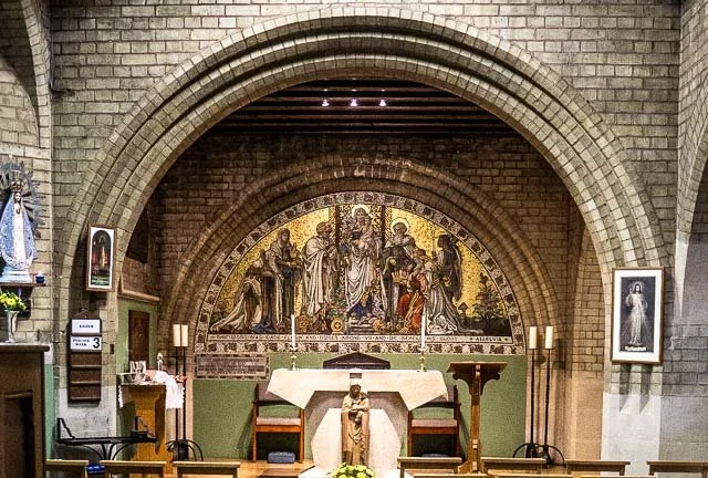 Inside a church, an altar with a white cloth, two candlesticks, and a statue at the front. Behind it is a large religious mosaic depicting Jesus Christ in the center, flanked by other figures. On the sides, framed religious photographs hang on the wa
