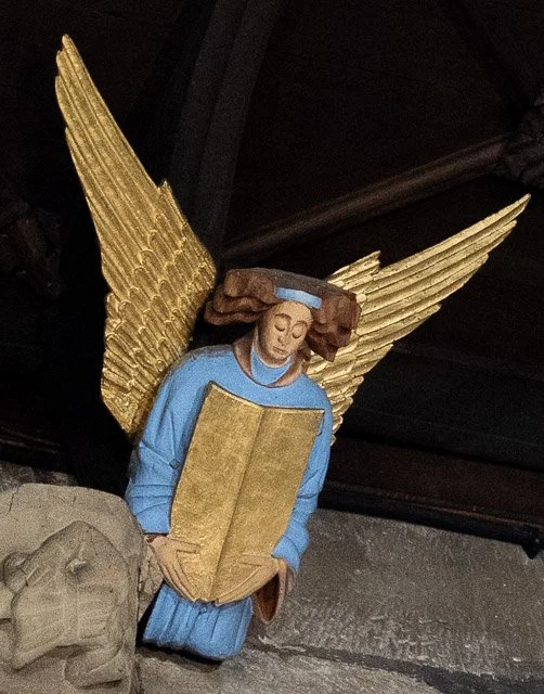 A carved wooden angel statue with golden wings, blue robe, brown hair, and holding an open book.