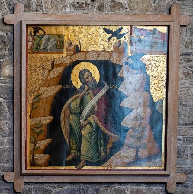 A religious icon painting depicting a saint with a halo, holding a scroll and a staff, surrounded by gold accents and trees, with a natural stone wall background.