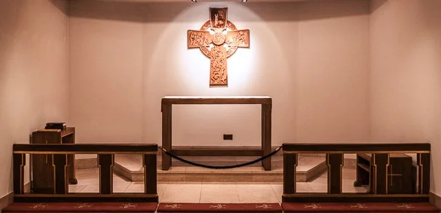 An empty church altar with a large wooden cross mounted on the back wall, illuminated by overhead lighting.