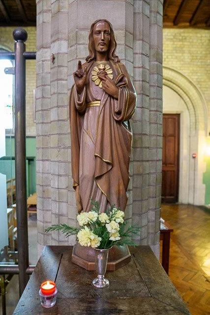 Wooden statue of Jesus Christ with one hand raised in blessing, holding a heart with a flame, in a church with a brick pillar and wooden door behind, flower arrangement and candle on table in front.