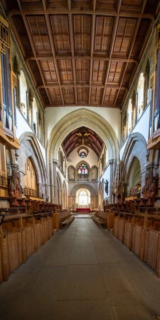 Interior of a historic church with wooden pews lining both sides, a vaulted ceiling, stained glass windows, and arched architecture at the front altar area.