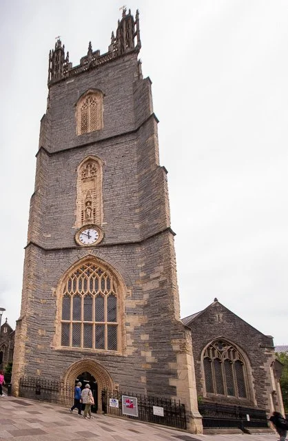 A tall, historic stone church tower with Gothic windows, a clock, and decorative battlements at the top, under an overcast sky, with people walking nearby.