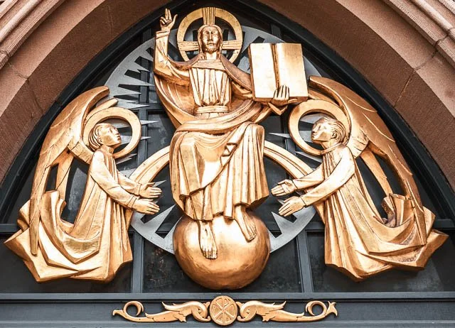 Gold-colored religious sculpture featuring Jesus Christ seated on a globe, holding an open book and raising his right hand in blessing. Two angels stand below, reaching up toward him. The sculpture is mounted on a black background with decorative ele