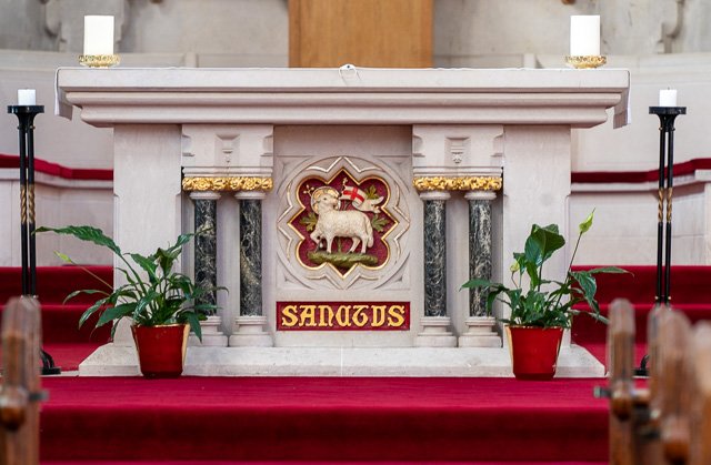 The altar of a church with an ornate white marble design, featuring the lamb of God emblem in the center, flanked by two potted plants, candles, and red carpeting.