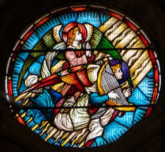 Stained glass window depicting a religious scene with an angel playing a harp and a person kneeling, surrounded by colorful geometric patterns.