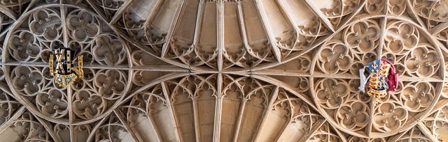 Close-up of ornate cathedral ceiling with intricate stone tracery and colorful royal coats of arms.