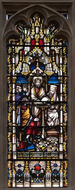 Stained glass window depicting a religious scene with a central figure, Jesus, surrounded by other figures in biblical attire. The window features intricate designs and vibrant colors.