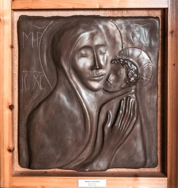 A bronze relief sculpture depicting a mother holding and gently touching her child, with religious symbols and inscriptions nearby.