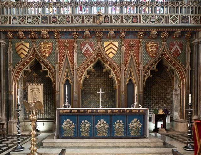 Interior view of a church altar with an ornate Gothic design, featuring a blue altar cloth with floral embroidery, a silver cross, and two candlesticks.