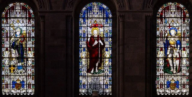 Three stained glass windows depicting religious figures inside a church, with dark stone framing.