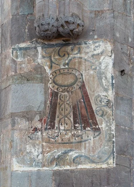 Ancient fresco painting of a shielded sword and a shield on a stone wall with decorative architectural element above.