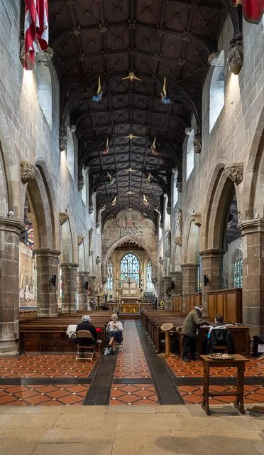 Interior of a church with high vaulted ceilings, stone walls, and stained glass windows at the end. Several people sit and stand at pews and tables along the sides.