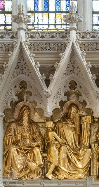 Gold religious sculptures of three figures, likely saints or religious figures, with detailed architectural white stone and stained glass windows above.