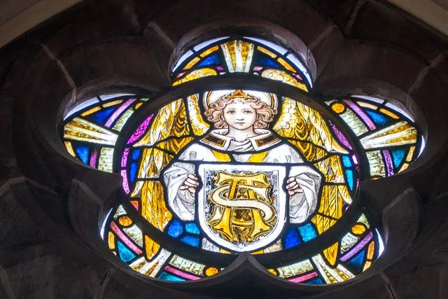 Stained glass window depicting an angel holding a shield with a golden symbol, surrounded by colorful patterns.