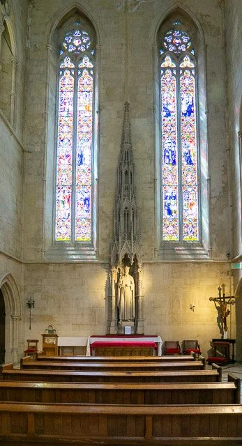 Inside a church with tall, stained glass windows, an altar with a red cloth, and wooden pews.