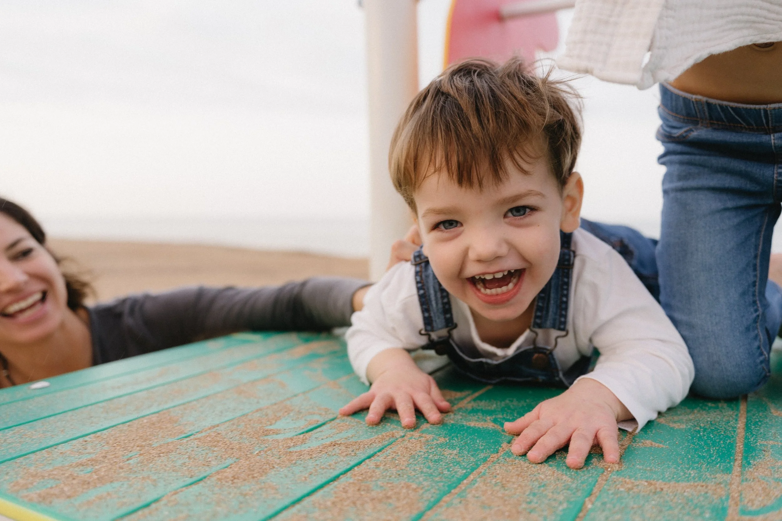 A smiling young boy with brown hair and blue eyes crawling on a sandy and green wooden surface, with an adult woman in the background at the beach.